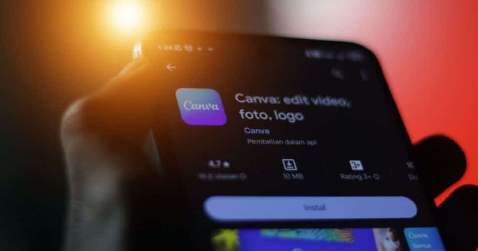 Brasil. Close-up de uma mão segurando um smartphone exibindo a página de instalação do aplicativo Canva, com informações como tamanho do app, classificação e opção para instalar. Fundo com iluminação em tons de laranja e vermelho