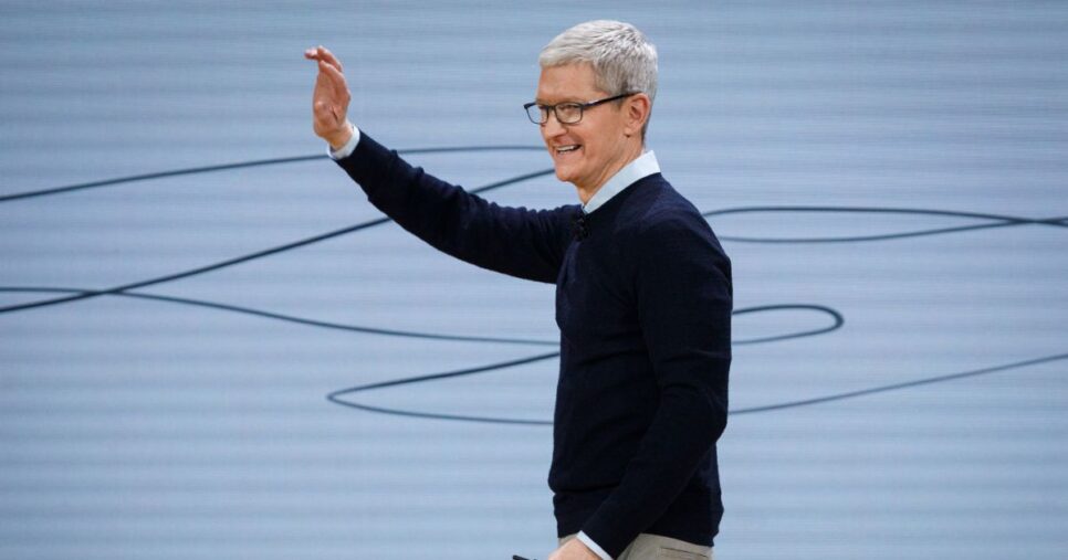 A cena mostra uma pessoa em um palco ou ambiente de apresentação, aparentemente durante um evento ou conferência tecnológica. O fundo é claro, com linhas curvas pretas que parecem fazer parte de um design minimalista. (Apple)