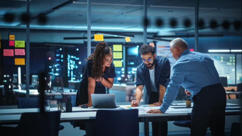 Três profissionais trabalhando juntos em uma reunião de negócios em um ambiente de escritório moderno, com laptops e quadros de planejamento ao fundo (nove