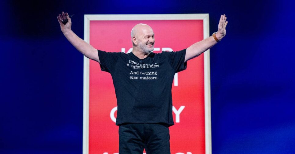 Homem calvo identificado como Werner Vogels, vice-presidente e CTO da Amazon, em pé no palco de um evento, sorrindo com os braços abertos; ele usa camiseta preta com frase em inglês e há um painel vermelho desfocado ao fundo