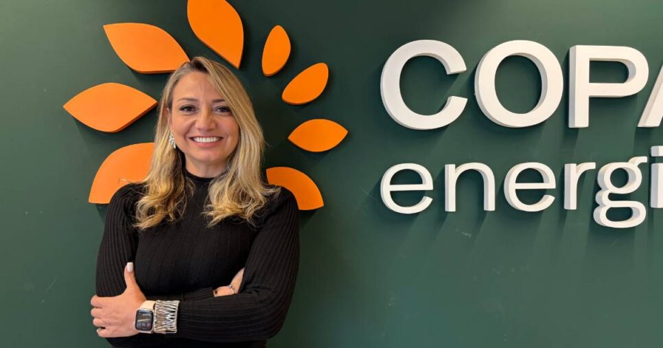 Mulher loira sorridente, de braços cruzados, usando blusa preta de manga comprida e relógio no pulso, posando em frente à parede verde com o logotipo da Copa Energia. Trata-se de Claudia Marquesani, CIO da empresa.