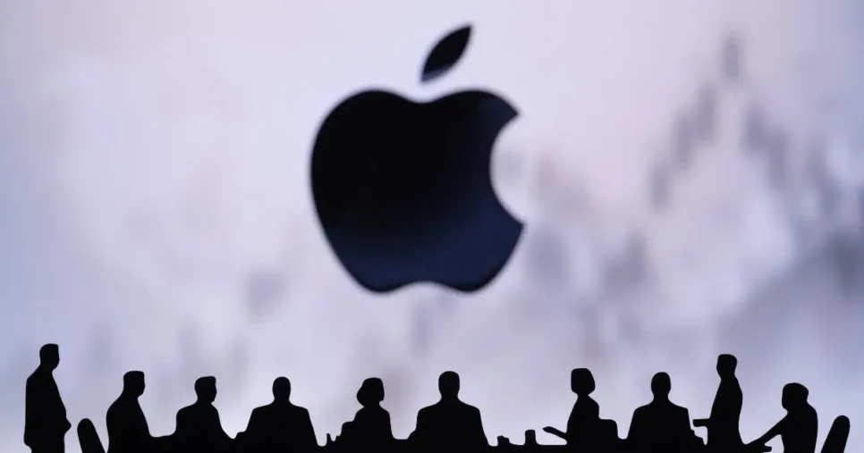 Silhuetas de pessoas sentadas à volta de uma mesa de reunião, em frente ao logótipo da Apple em destaque ao fundo, com um gráfico desfocado ao fundo sugerindo contexto financeiro ou empresarial.
