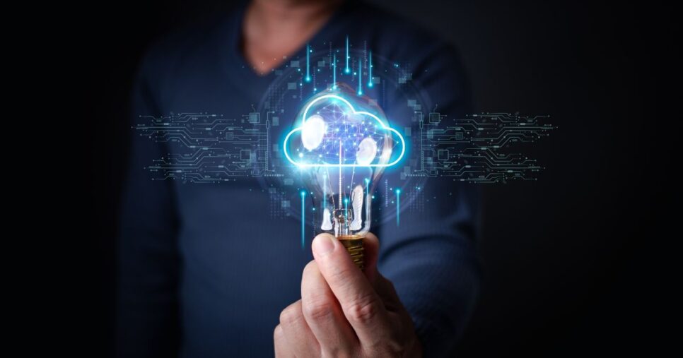 Uma pessoa segura uma lâmpada acesa, da qual emerge um holograma em forma de nuvem com padrões que lembram circuitos eletrônicos. O fundo é escuro, o que destaca os elementos brilhantes em azul e branco do holograma. (Seidor, nuvem, transforma)