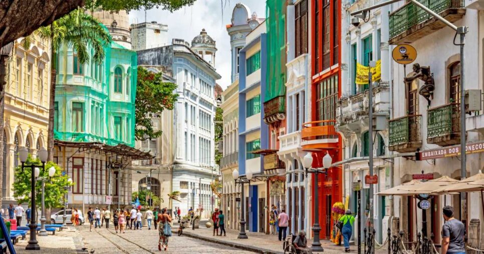 Rua do bairro do Recife Antigo, em Pernambuco, com fachadas coloridas e históricas, pessoas caminhando pela via de paralelepípedos e clima ensolarado. (Simpress)