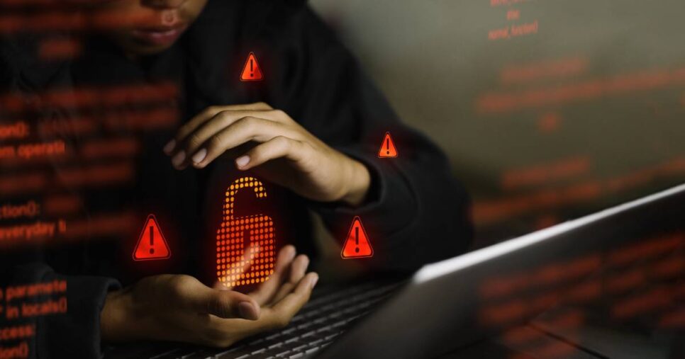 Pessoa parcialmente visível usando laptop no escuro, com as mãos em volta de um ícone digital de cadeado aberto iluminado em vermelho, cercado por símbolos de alerta, representando vazamento de dados ou falha de segurança
