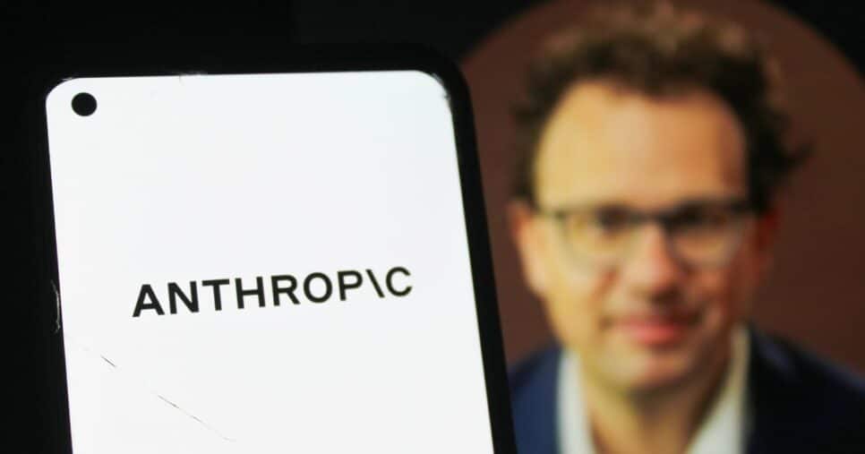 Telefone celular exibindo, em destaque, o nome “ANTHROPIC” em letras pretas sobre fundo branco. Ao fundo, aparece uma pessoa usando roupas formais, em segundo plano, com iluminação suave e foco concentrado no dispositivo móvel.