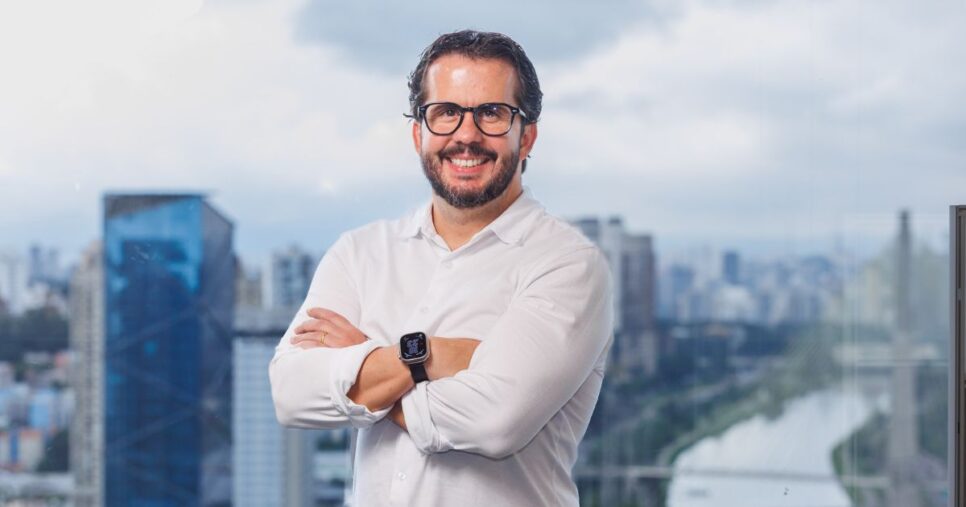 A imagem mostra uma pessoa em pé, com os braços cruzados, em frente a uma grande janela panorâmica que revela uma vista urbana. O cenário sugere um ambiente corporativo ou profissional. (Cohesity)