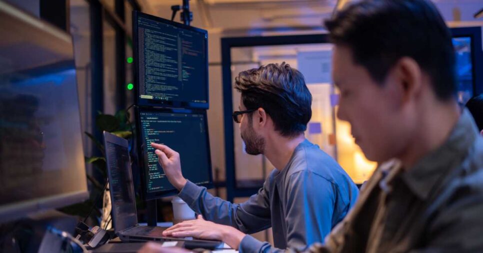 Dois desenvolvedores trabalham lado a lado em um escritório moderno, olhando para múltiplos monitores com códigos na tela; um deles aponta para o monitor enquanto o outro digita no notebook, vibe coding, proteção, 2026
