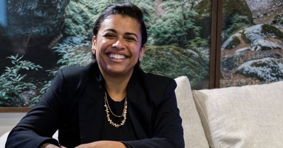 Mulher sorridente sentada em um sofá claro, usando blazer preto e colar dourado, com um painel de paisagem natural ao fundo; junto à imagem aparece o texto “Noéle Gomes, pesquisadora”. (filosofia)