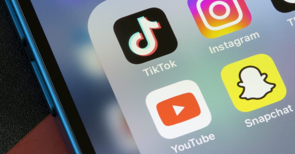 Imagem mostra a tela de um smartphone com os ícones dos aplicativos TikTok, Snapchat, Instagram e YouTube organizados em uma pasta. O aparelho possui borda azul metálica e está sobre uma superfície escura.(União Europeia, Kaspersky)