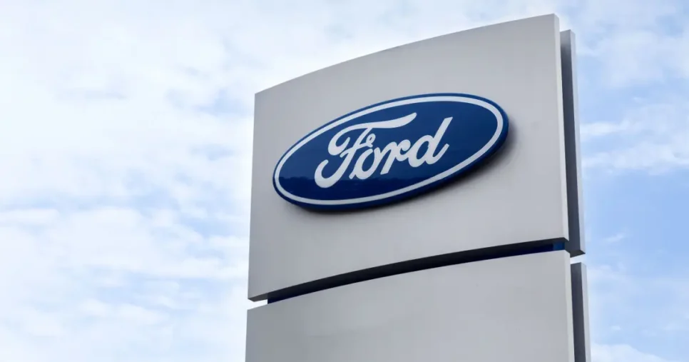 Imagem de um grande letreiro da Ford, com o logotipo oval azul e a palavra "Ford" escrita em fonte cursiva branca. O letreiro está fixado em uma estrutura metálica cinza clara, com o céu parcialmente nublado ao fundo (agendamentos)