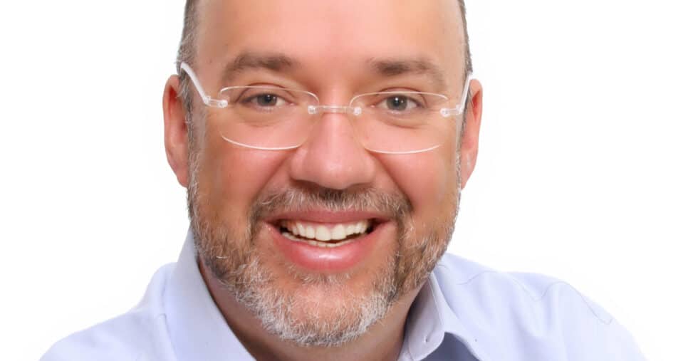 Jorge Luís Cordenonsi, CIO da Compass Latin America e GRSA, sorrindo, usando óculos de armação transparente, camisa azul clara e fundo branco.