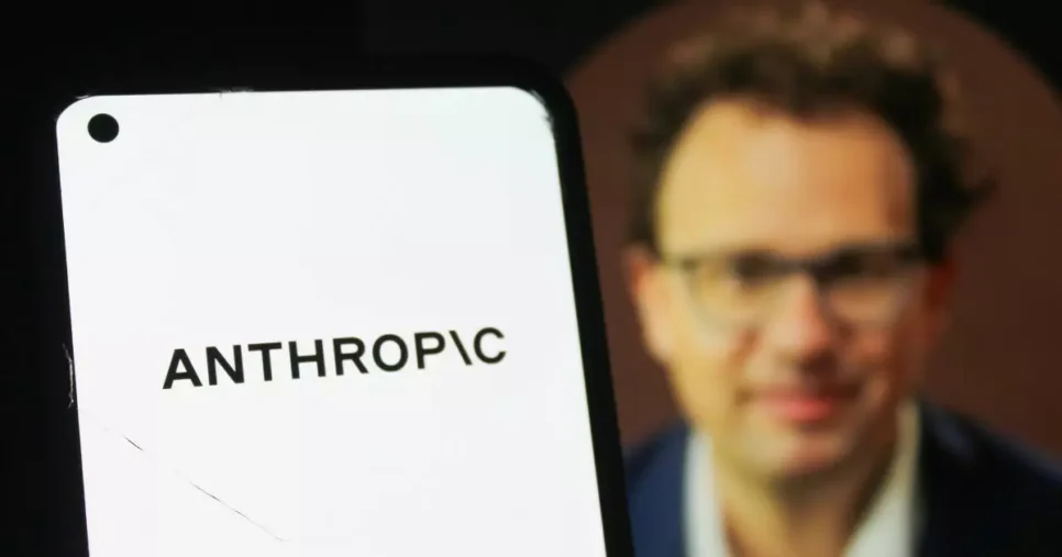 Telefone celular exibindo, em destaque, o nome “ANTHROPIC” em letras pretas sobre fundo branco. Ao fundo, aparece uma pessoa usando roupas formais, em segundo plano, com iluminação suave e foco concentrado no dispositivo móvel. (humanidade)