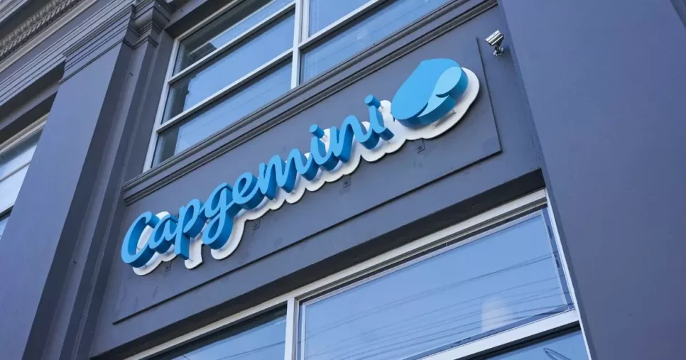 A imagem mostra a parte externa de um edifício com uma placa que exibe o nome "Capgemini" em letras cursivas azuis, acompanhadas de um logotipo azul e branco. A placa está fixada na fachada do prédio, que possui grandes janelas. (tendencias)