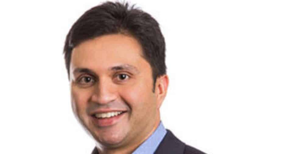 A imagem mostra uma pessoa vestindo um blazer escuro sobre uma camisa social azul‑clara, posicionada diante de um fundo totalmente branco. A composição é simples e focada na parte superior do corpo, com iluminação uniforme. (Netskope). Forescout