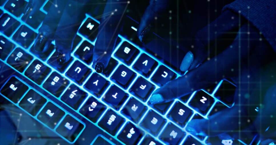 Simulação de cibercrime com imagem em close-up de mãos digitando em um teclado retroiluminado com luz azul. As teclas estão iluminadas e os dedos estão posicionados sobre diferentes teclas. (Anthropic, coding)