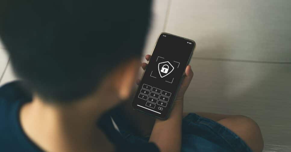 Uma criança sentada no chão segura um smartphone cuja tela exibe um cadeado e um teclado numérico de senha, sugerindo uma interface de bloqueio ou segurança digital. A cena é iluminada suavemente e o foco está no dispositivo.