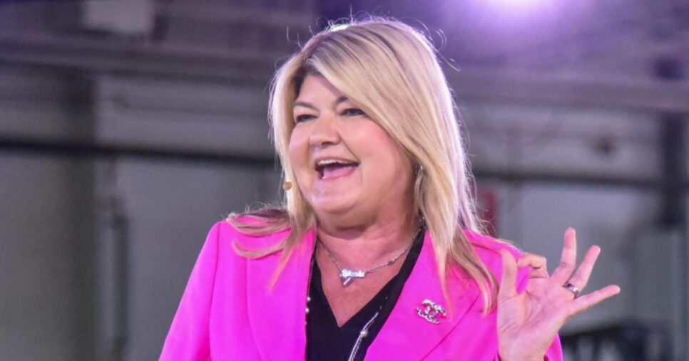 Pessoa em pé atrás de um púlpito transparente, vestindo um blazer rosa-choque sobre uma blusa preta. O rosto da pessoa está desfocado. Ela faz um gesto de 'OK' com a mão direita. (IA, sandy carter)