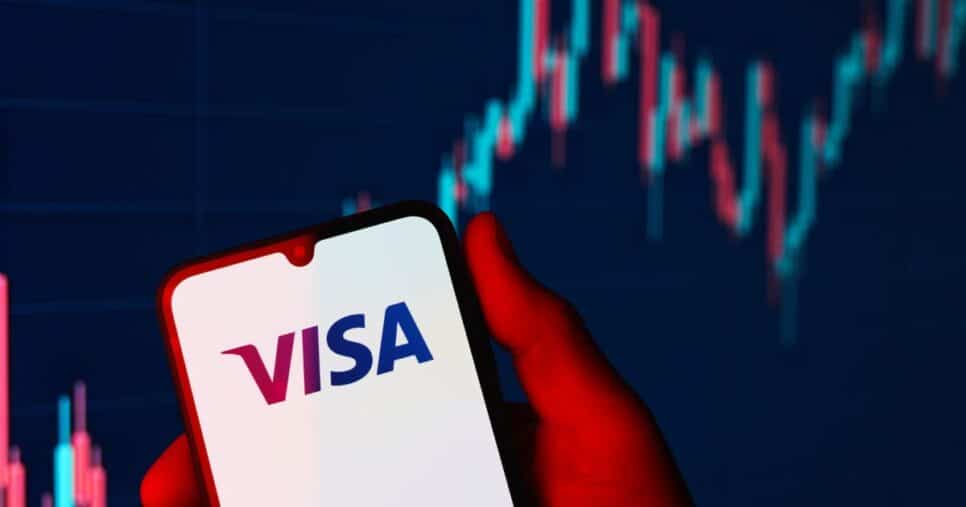 Uma mão segura um smartphone que exibe o logotipo da VISA na tela. Ao fundo, há um gráfico financeiro com padrões de velas (candlestick), representando dados do mercado de ações.