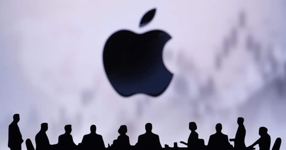 Silhuetas de pessoas sentadas à volta de uma mesa de reunião, em frente ao logótipo da Apple em destaque ao fundo, com um gráfico desfocado ao fundo sugerindo contexto financeiro ou empresarial.