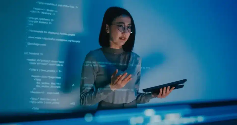 Mulher jovem usando óculos e segurando um tablet, trabalhando com códigos de programação em um ambiente com luz azul e tecnologia avançada.