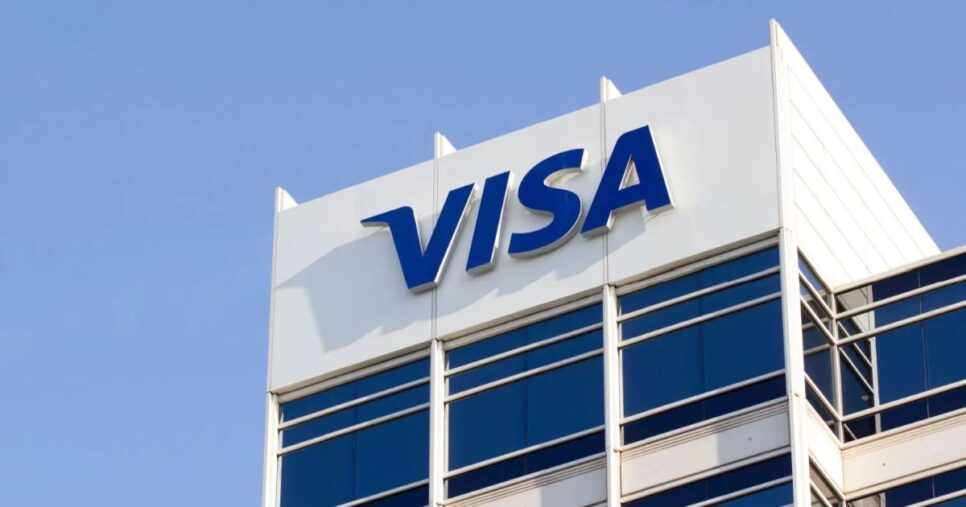 Fachada de um prédio corporativo da Visa com o logotipo azul da empresa em destaque sobre um fundo branco, contra um céu azul claro. A estrutura moderna do edifício é composta por janelas de vidro refletivo (visa, vai de visa)